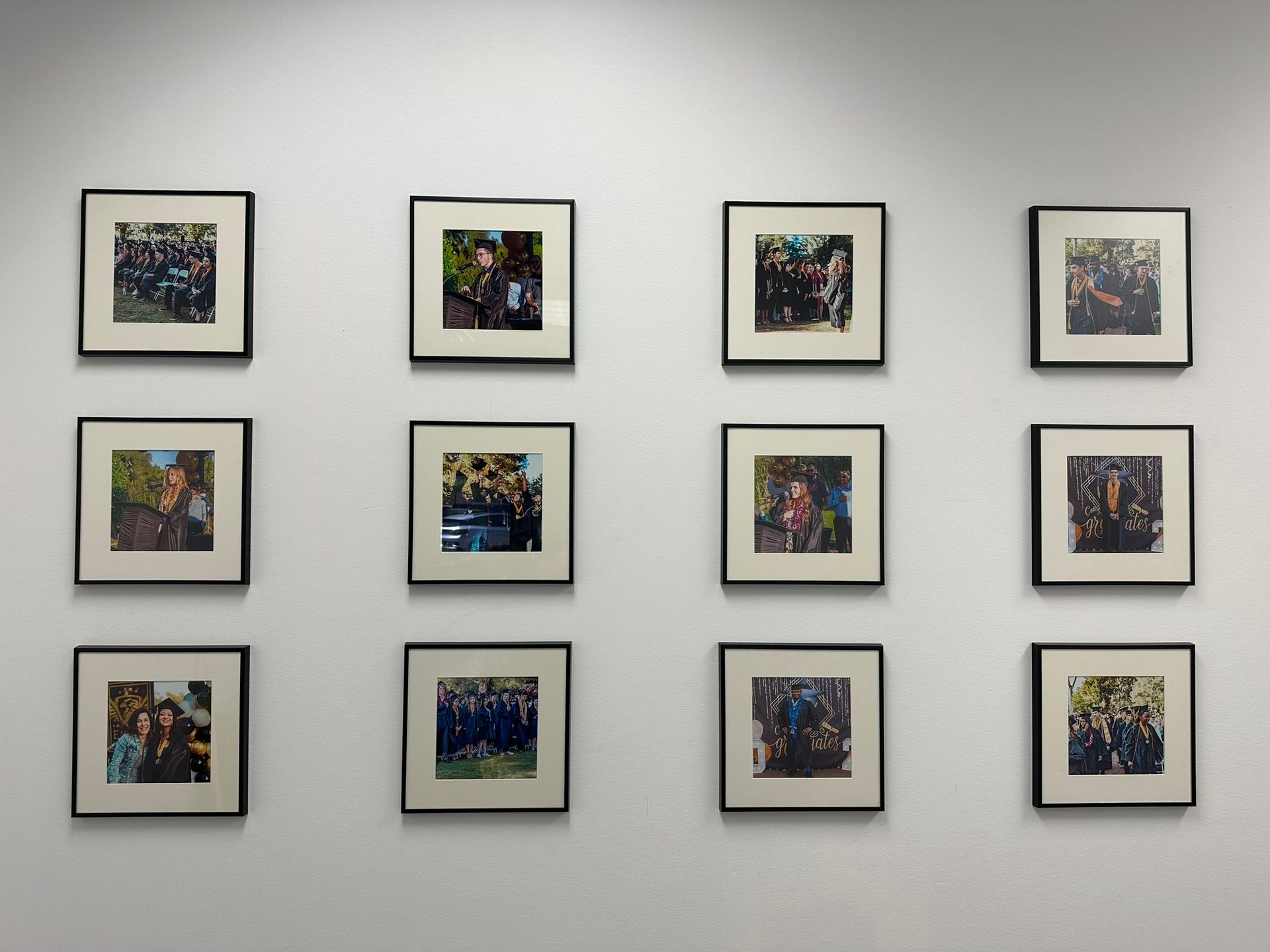 Twelve framed photos displayed on a white wall. Each black frame encloses a photograph of people.
