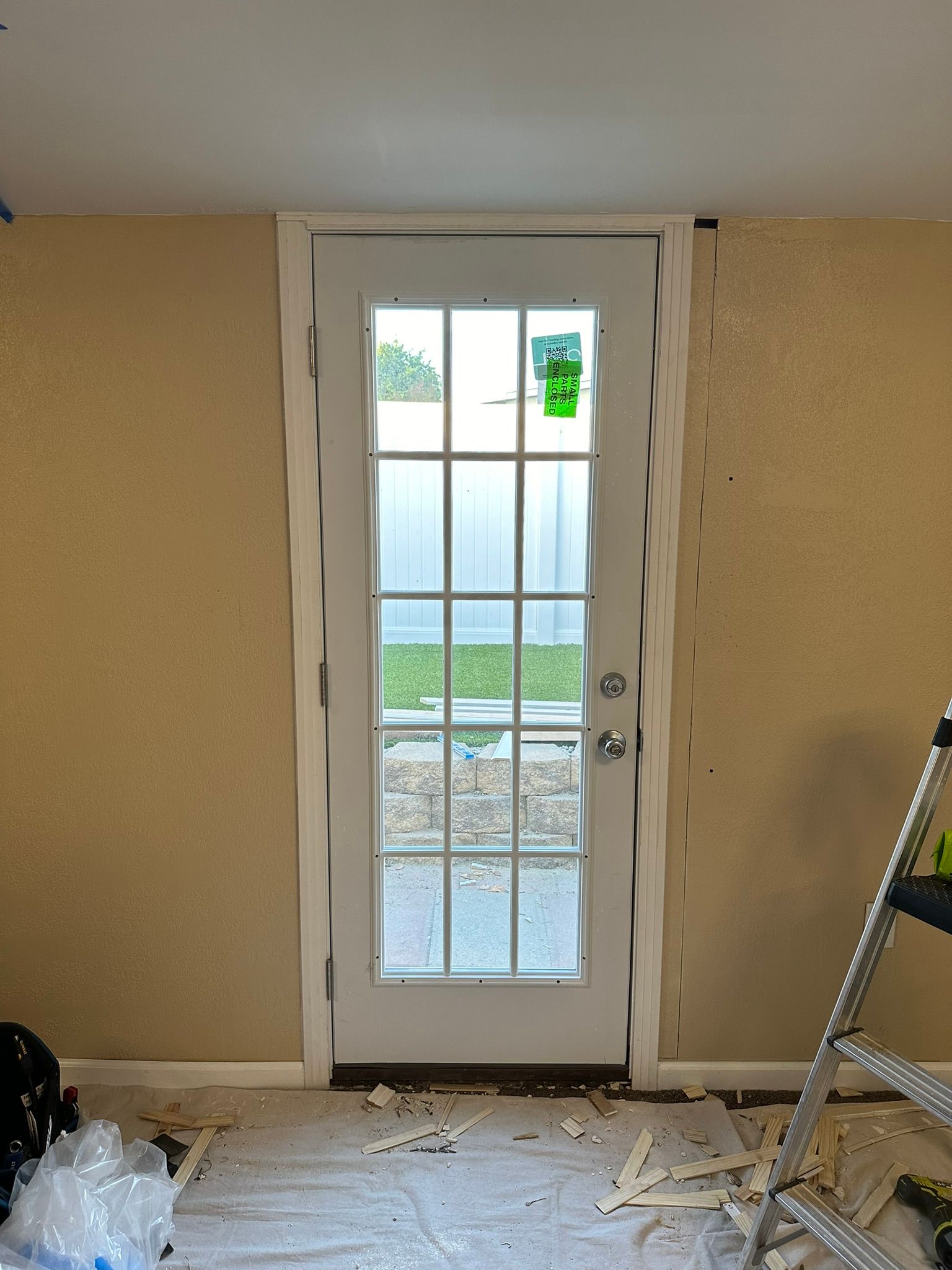 White French door in a tan-walled room, trim partially installed, yard visible through glass panes, ladder present.