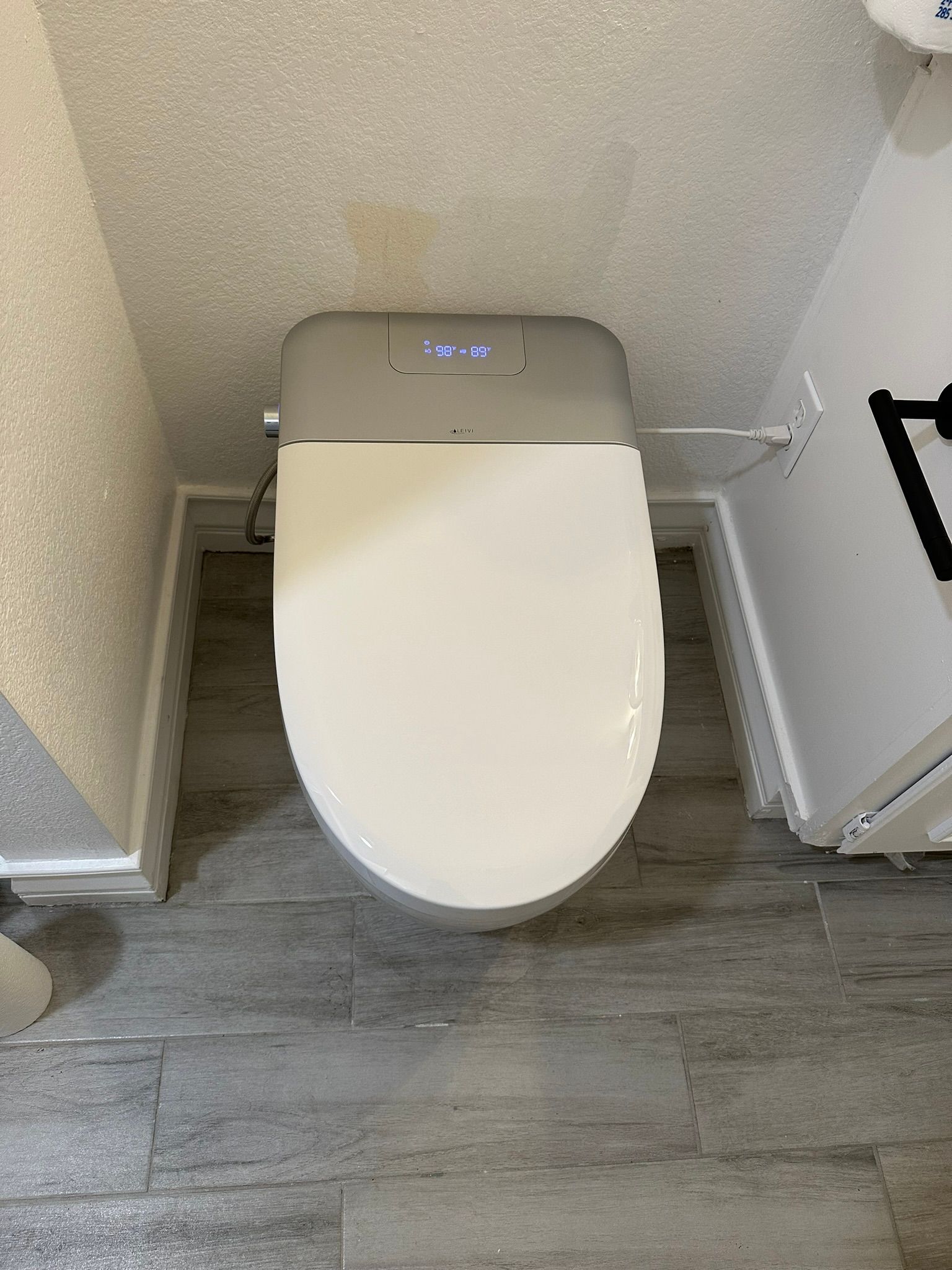 Top-down view of a white, elongated toilet with a gray electronic bidet seat in a small bathroom. Gray tiled floor and white walls.