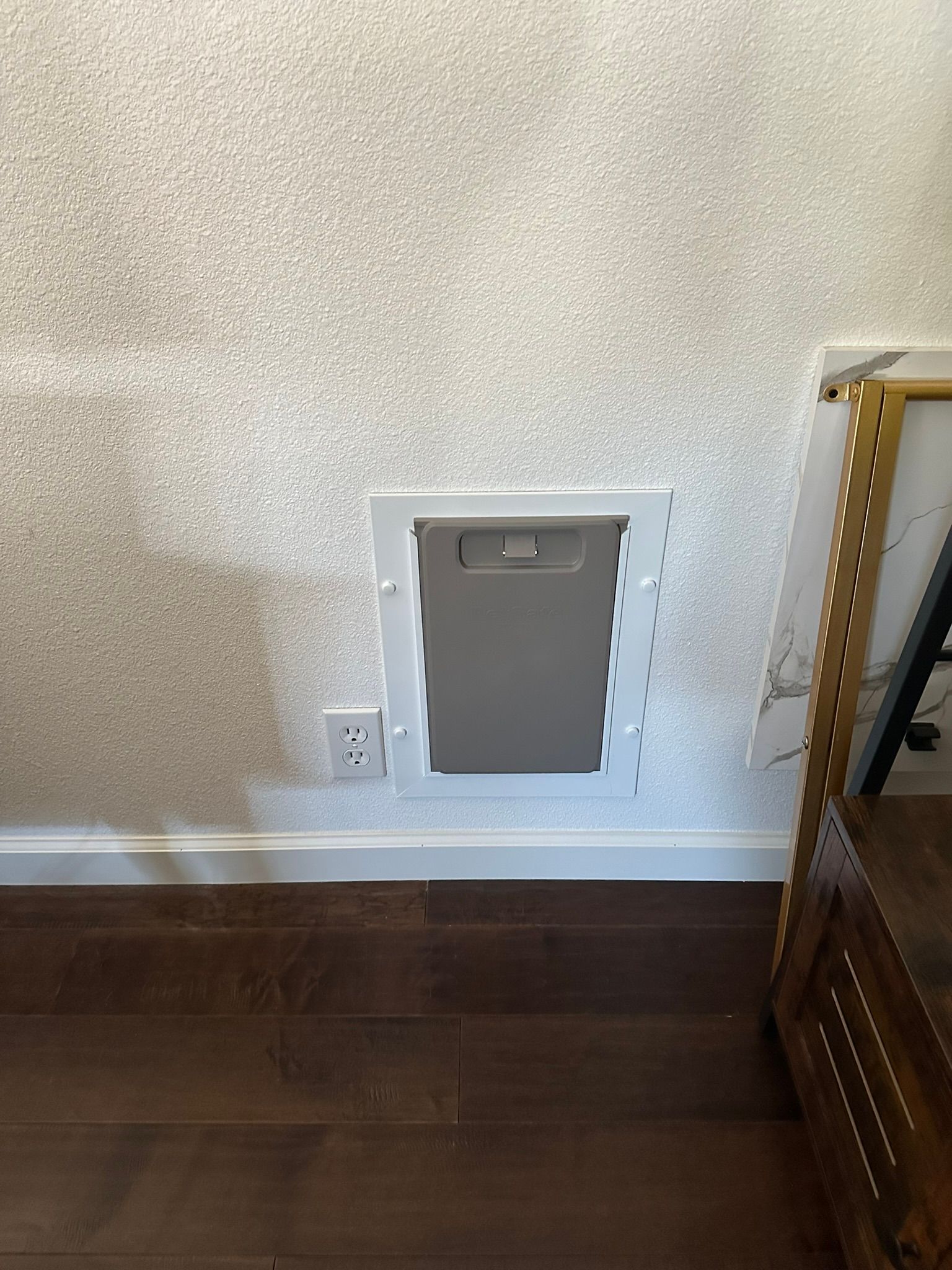 A wall-mounted access panel with a recessed door, next to a power outlet and a wooden floor.