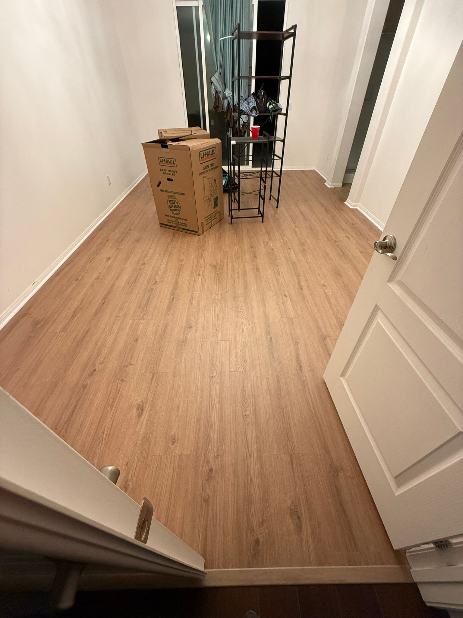 A long, narrow room with wood-look flooring. A tall shelf unit and cardboard box are in the space.