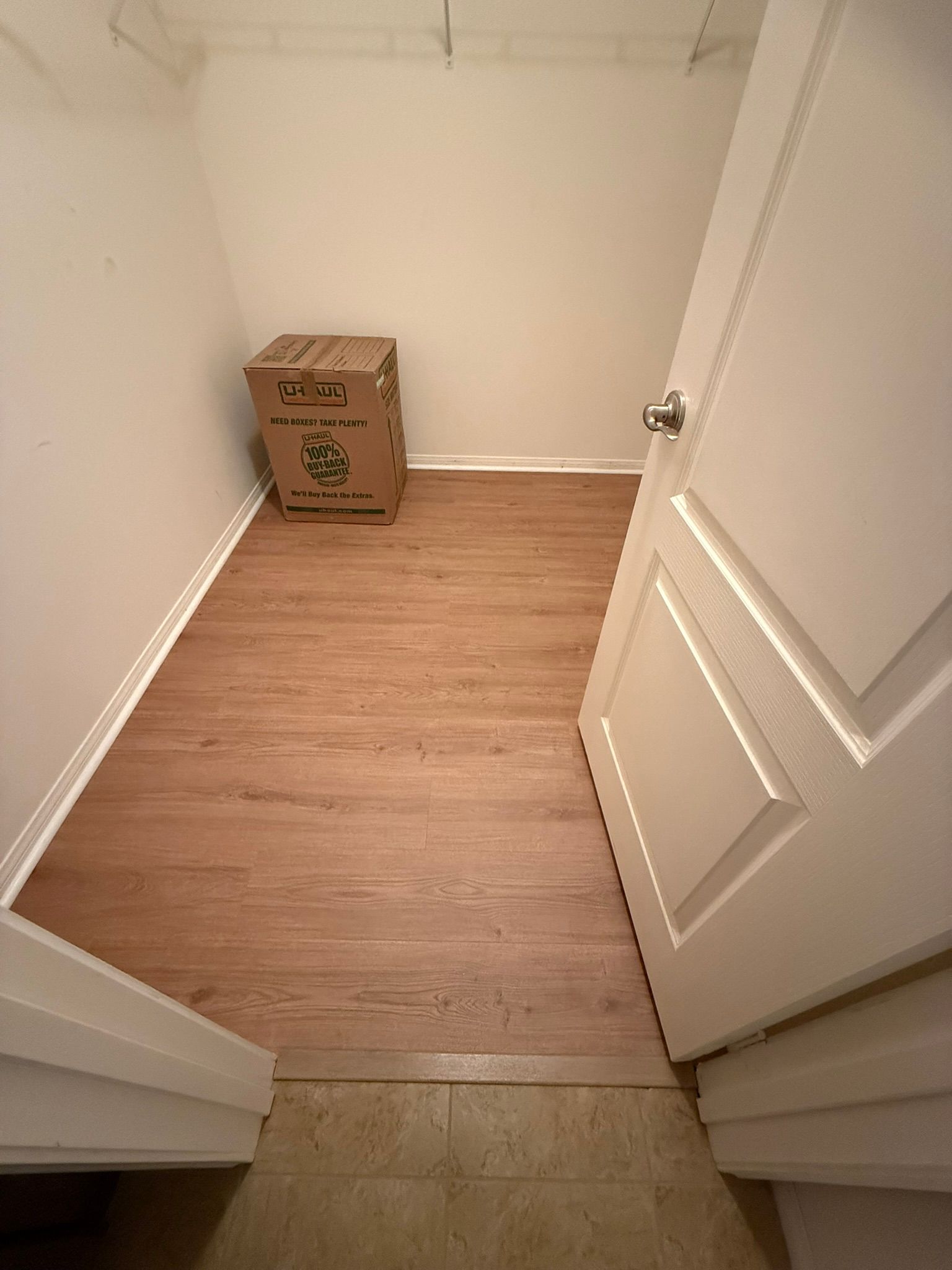 Small closet with wood-look flooring and a cardboard box. The door is ajar, revealing a tiled entrance.
