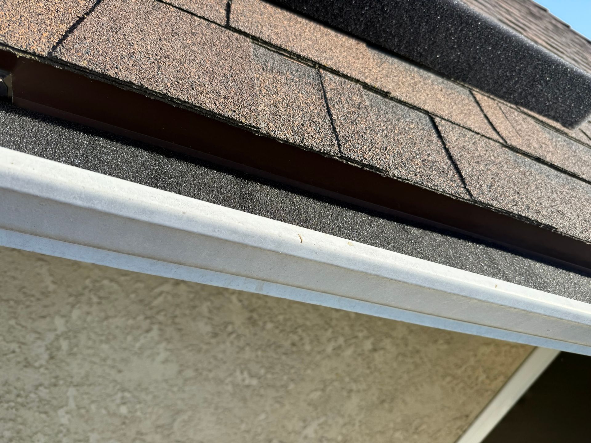 Close-up of a roof edge with brown shingles, a dark underlayment, and a white gutter.