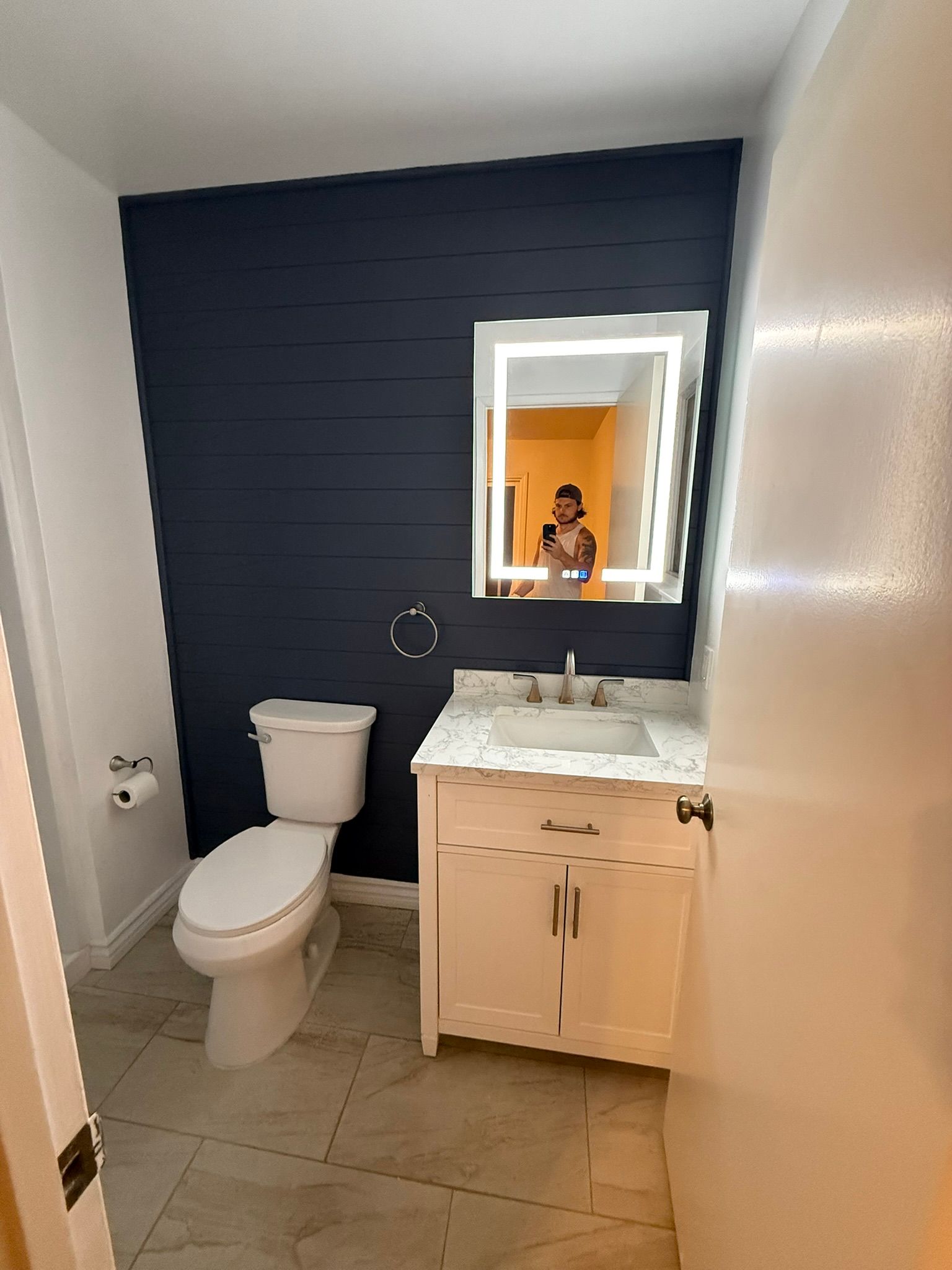 Bathroom with navy shiplap wall, toilet, white vanity, lit mirror, and light tiled floor.