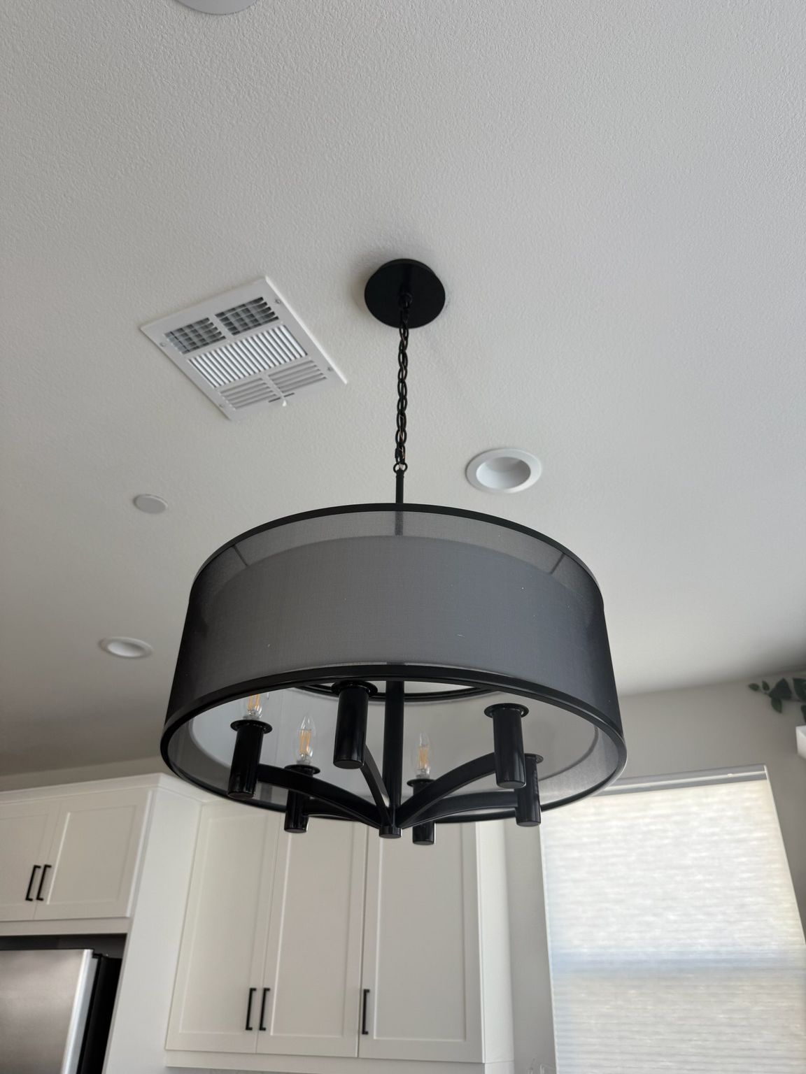 A modern black drum-style chandelier hangs from a white ceiling above a kitchen with white cabinetry.