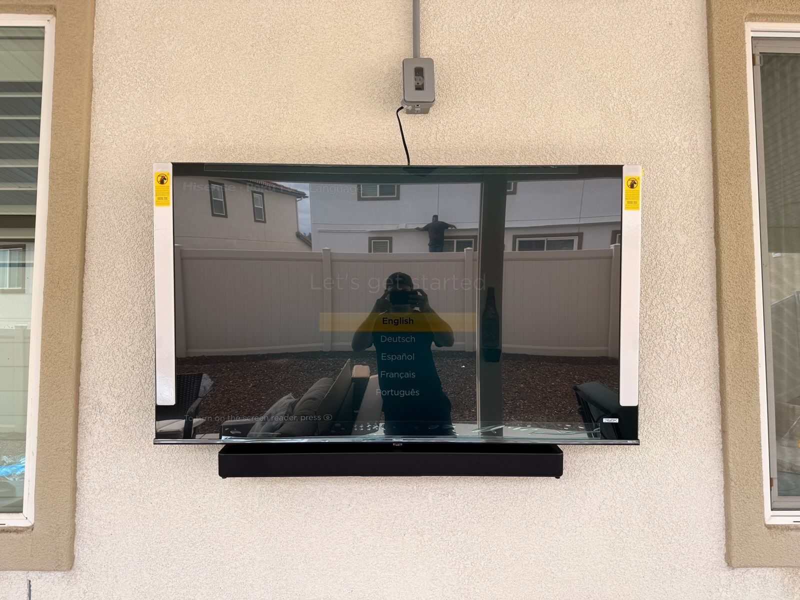 A flat-screen television with a soundbar is mounted on an exterior wall between two windows.