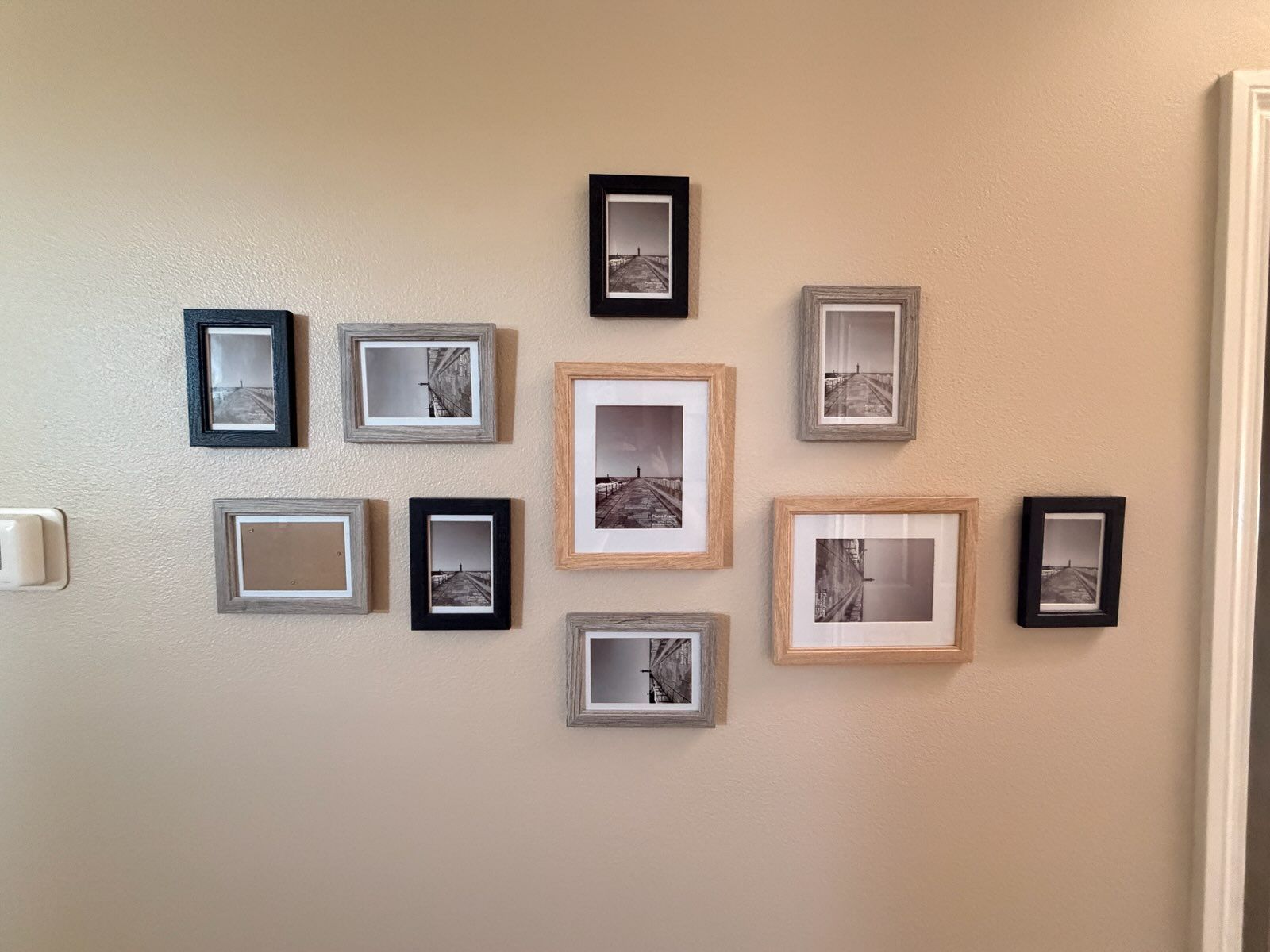 A gallery wall display featuring ten mixed-size black, grey, and light wood frames containing black and white photos.