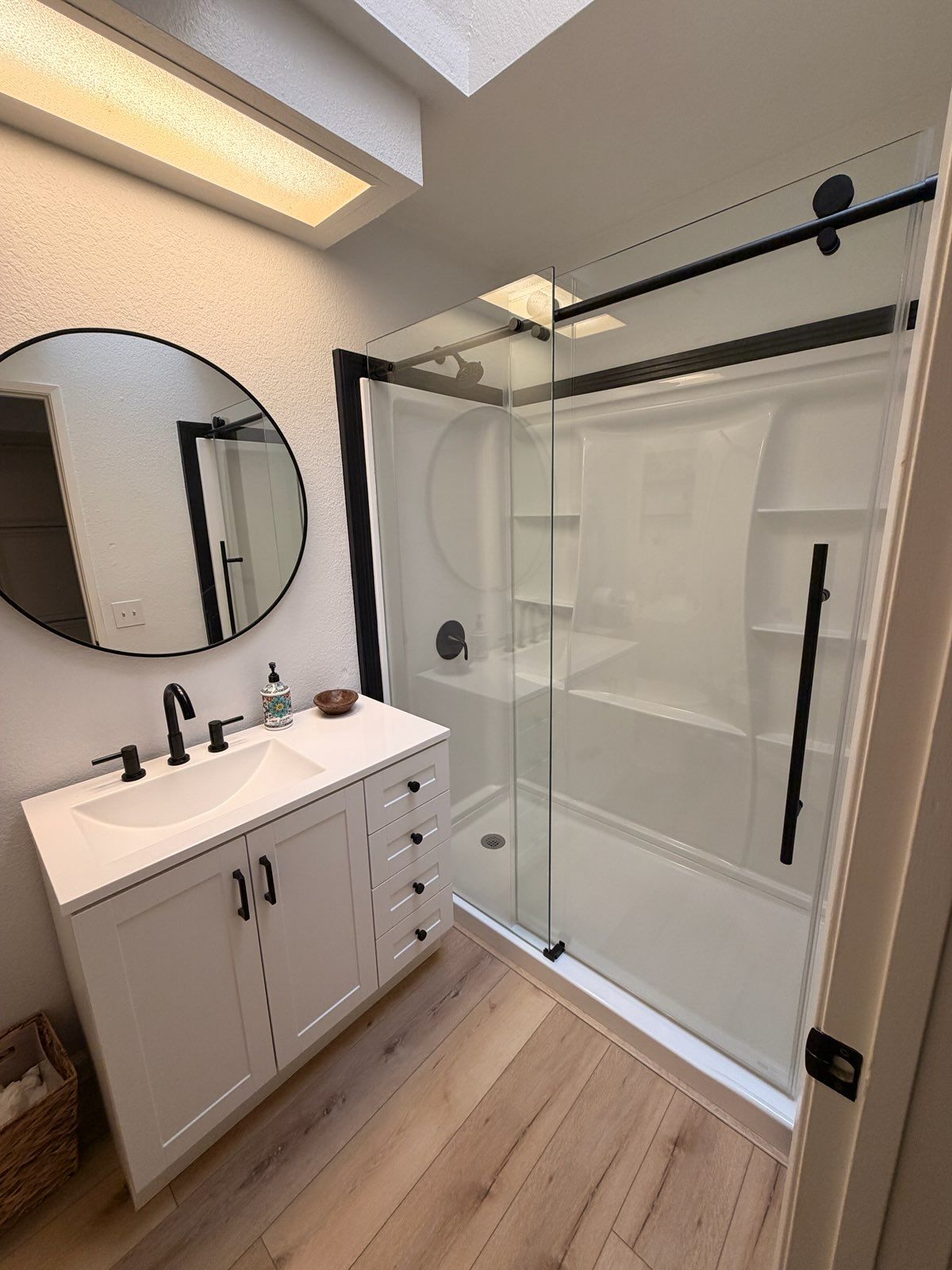 Modern bathroom with white vanity, round mirror, and a glass-enclosed walk-in shower with black hardware on wood floors.