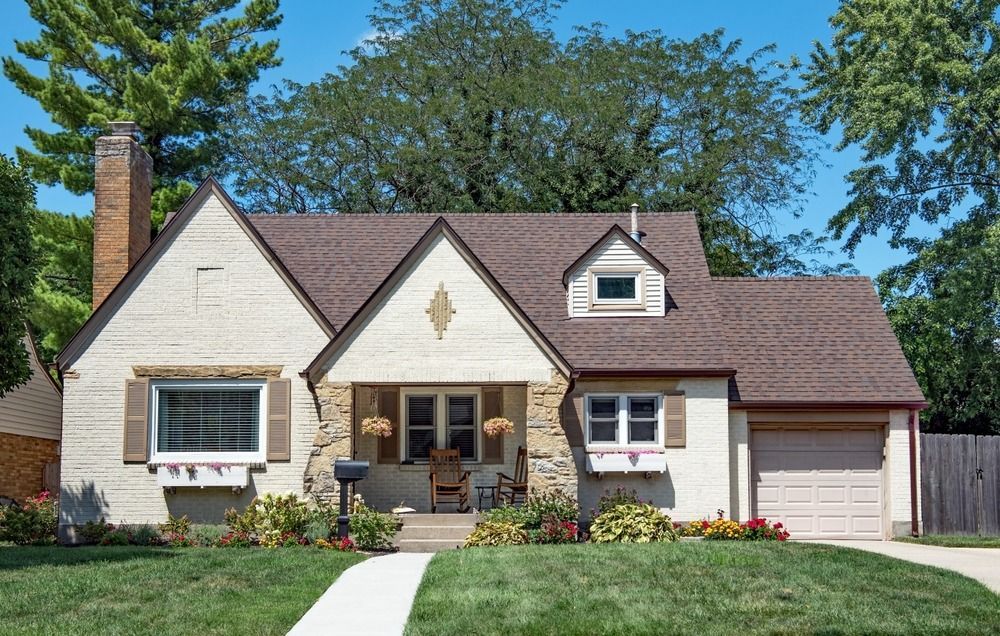 Cottage-style house with brown roof, tan exterior, and a small garage, set against a backdrop of green trees.