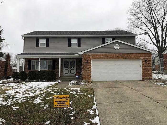 Two-story house with gray siding, brick garage, and front porch on a winter day with patchy snow.