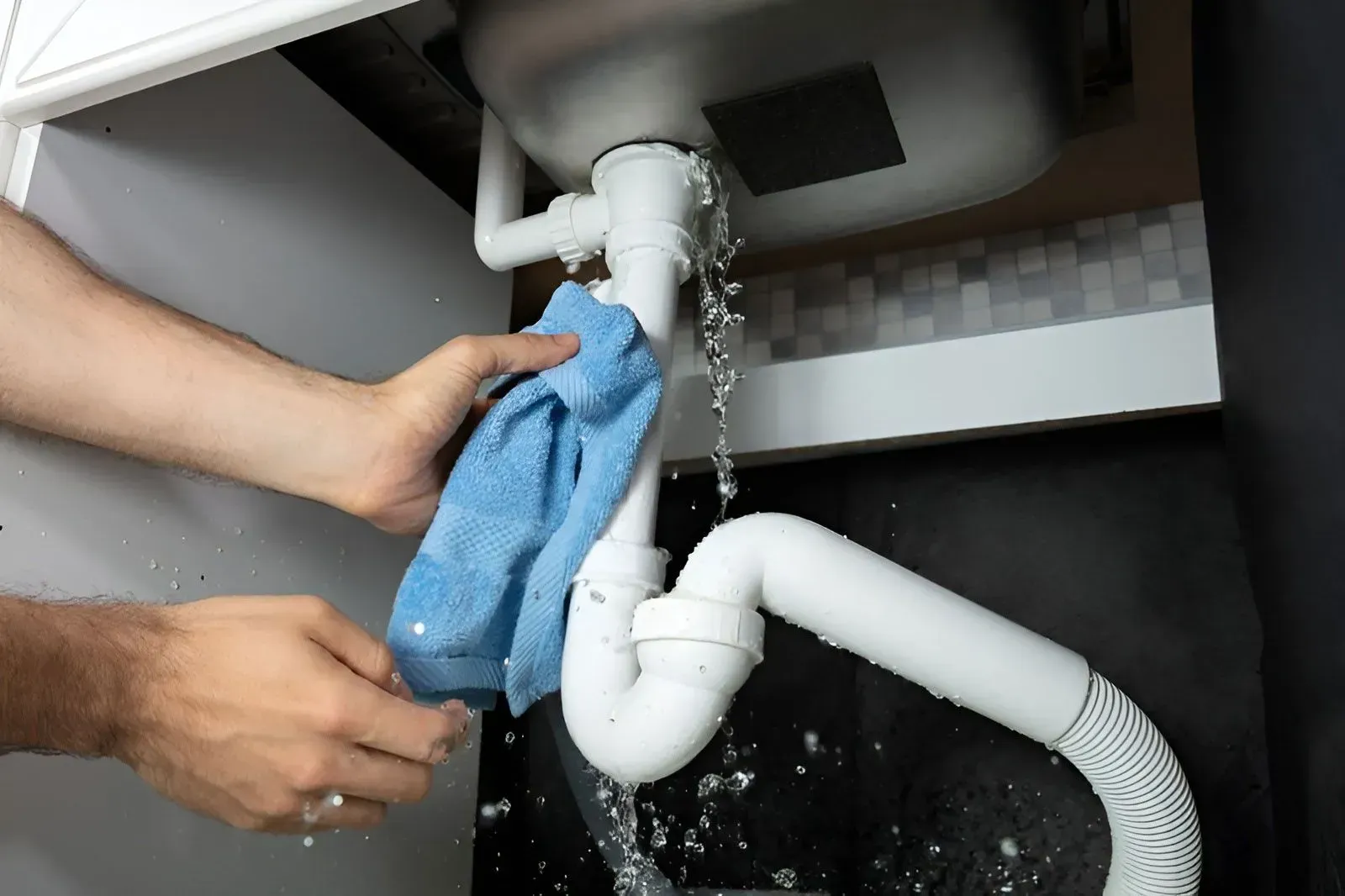 A Person Holds a Blue Cloth to a Leaking White Kitchen Sink Pipe — Dunnrite Plumbing Services in Airlie Beach, QLD