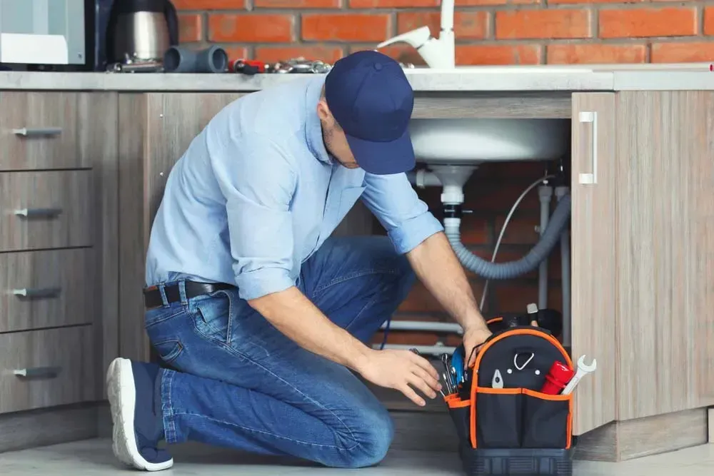 A Plumber in a Cap and Work Clothes Kneels While Reaching for Tools — Dunnrite Plumbing Services in Cannonvale, QLD
