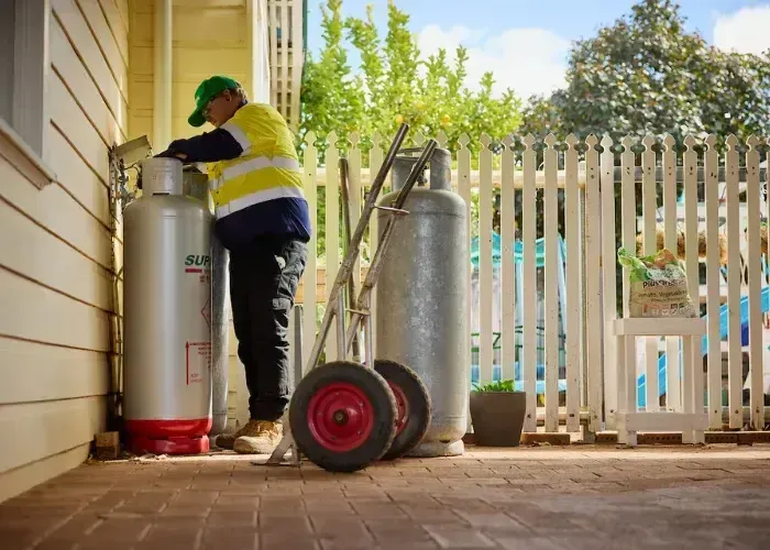 Worker checking a tall gas cylinder beside a house patio and fence - Dunnrite Plumbing Services in Cannonvale, QLD
