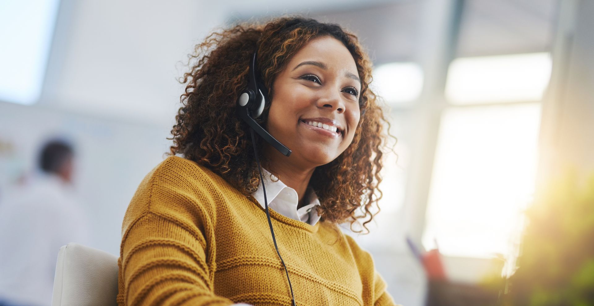 A woman is wearing a headset and smiling while sitting at a desk.