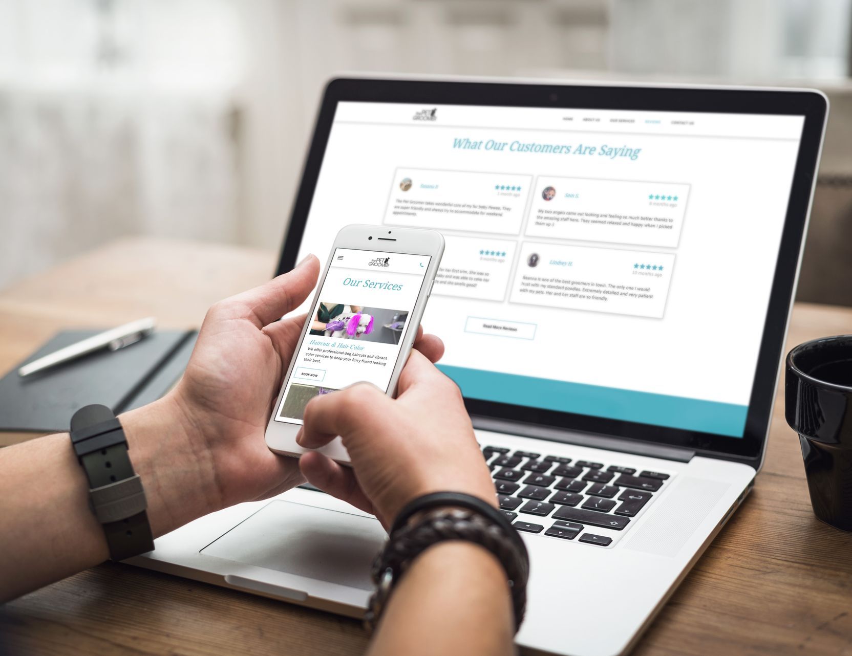 A person is holding a cell phone in front of a laptop computer viewing The Pet Groomer website