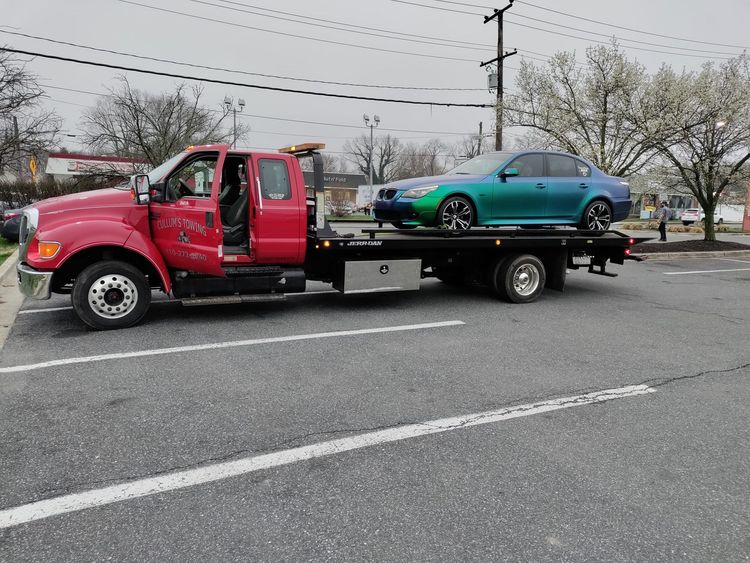 Red tow truck carrying a green hatchback on a wet roadside under gray skies