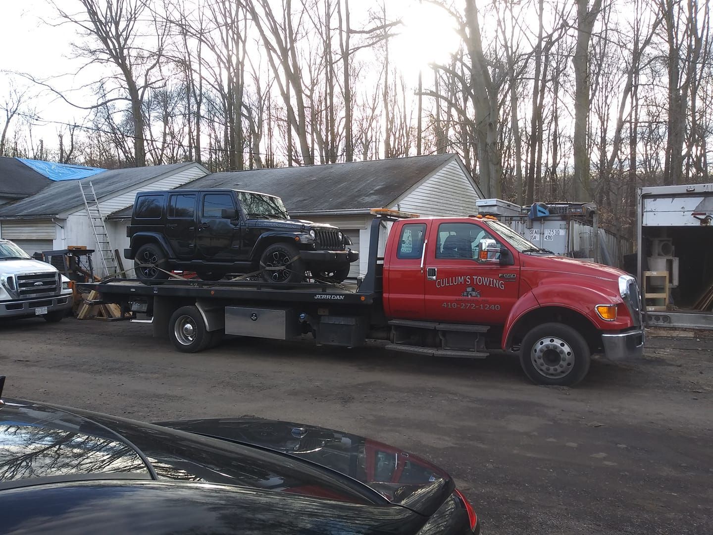 Red tow truck carrying a black SUV in a lot beside a low building and bare trees