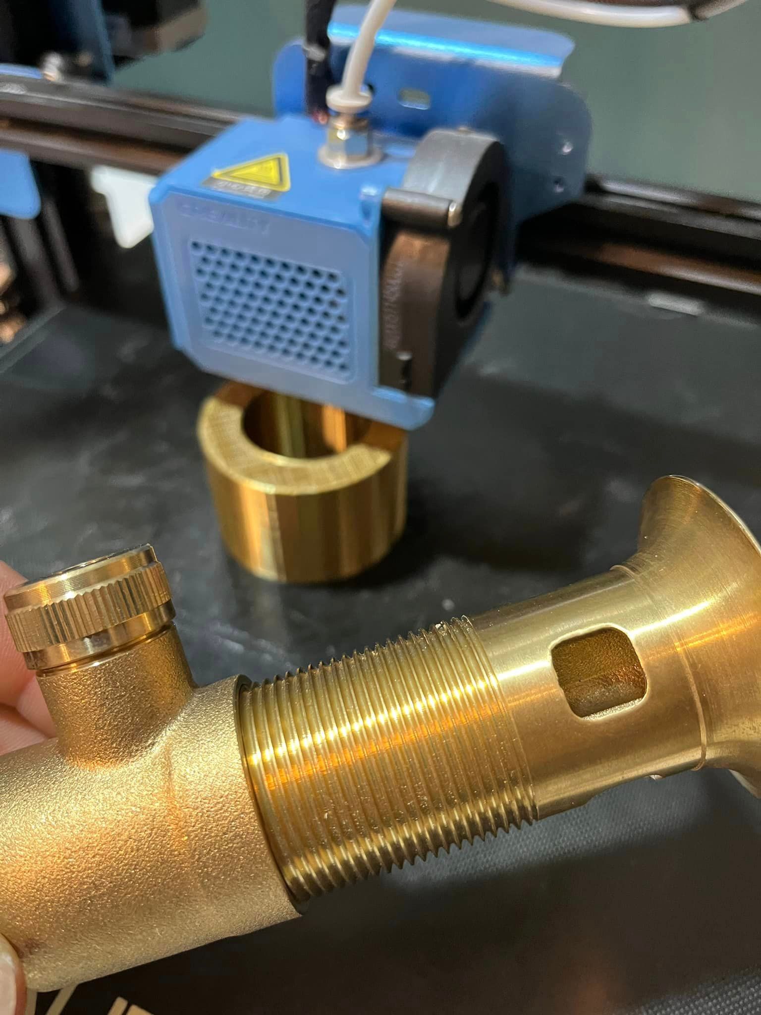 A person is holding a brass pipe in front of a 3d printer.