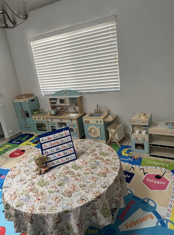 Child’s playroom with a round floral table, toy kitchen sets, and a window with white blinds.
