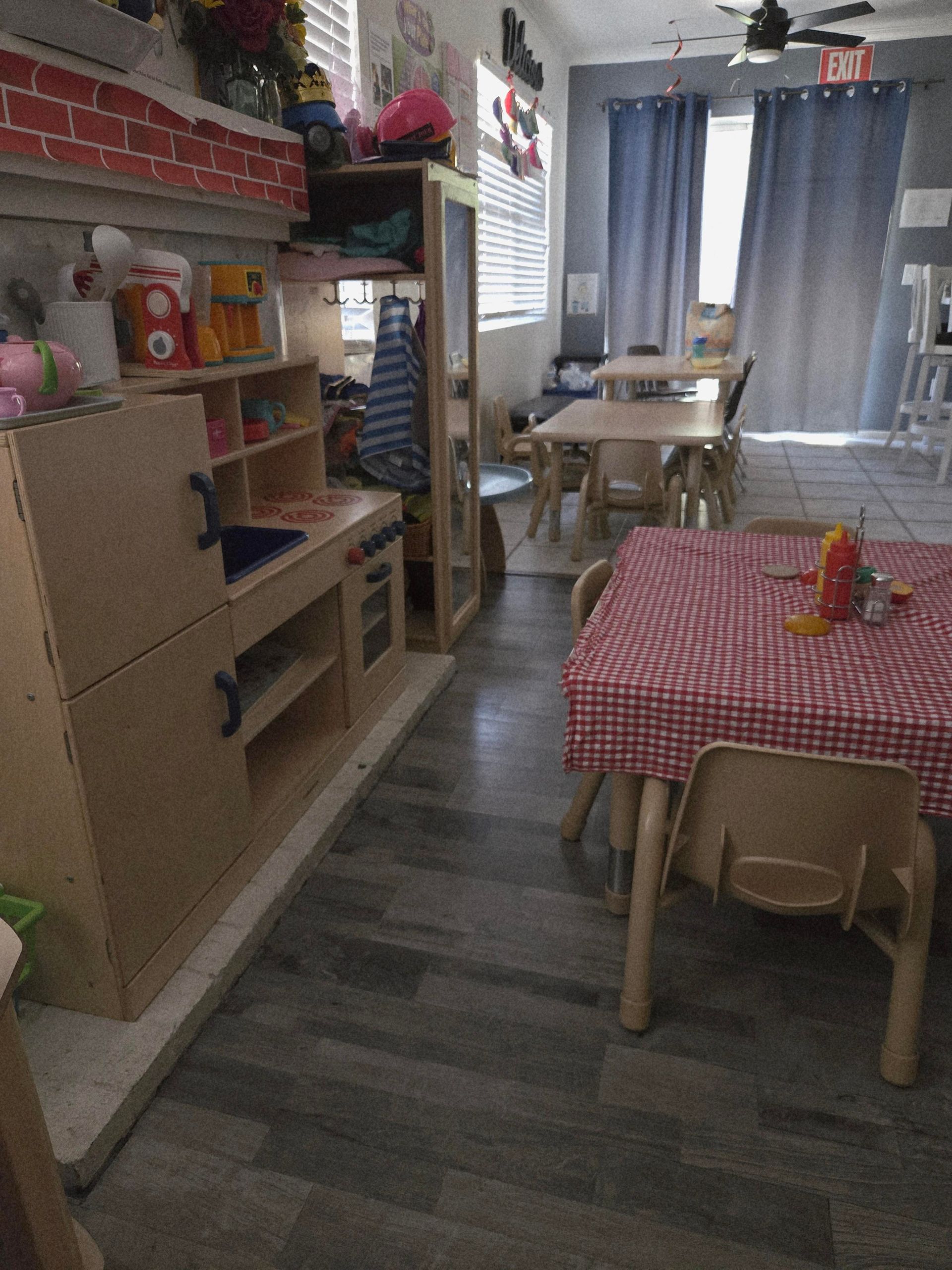 Small dining area with a checkered tablecloth, wooden chairs, shelving, and blue curtains at the back.