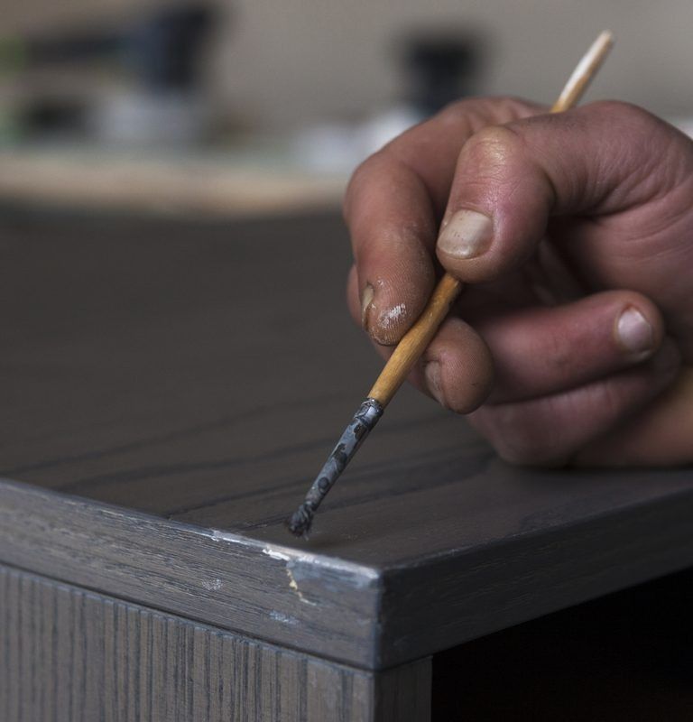 Mano che tiene un pennello, mentre dipinge una superficie di legno grigio scuro.