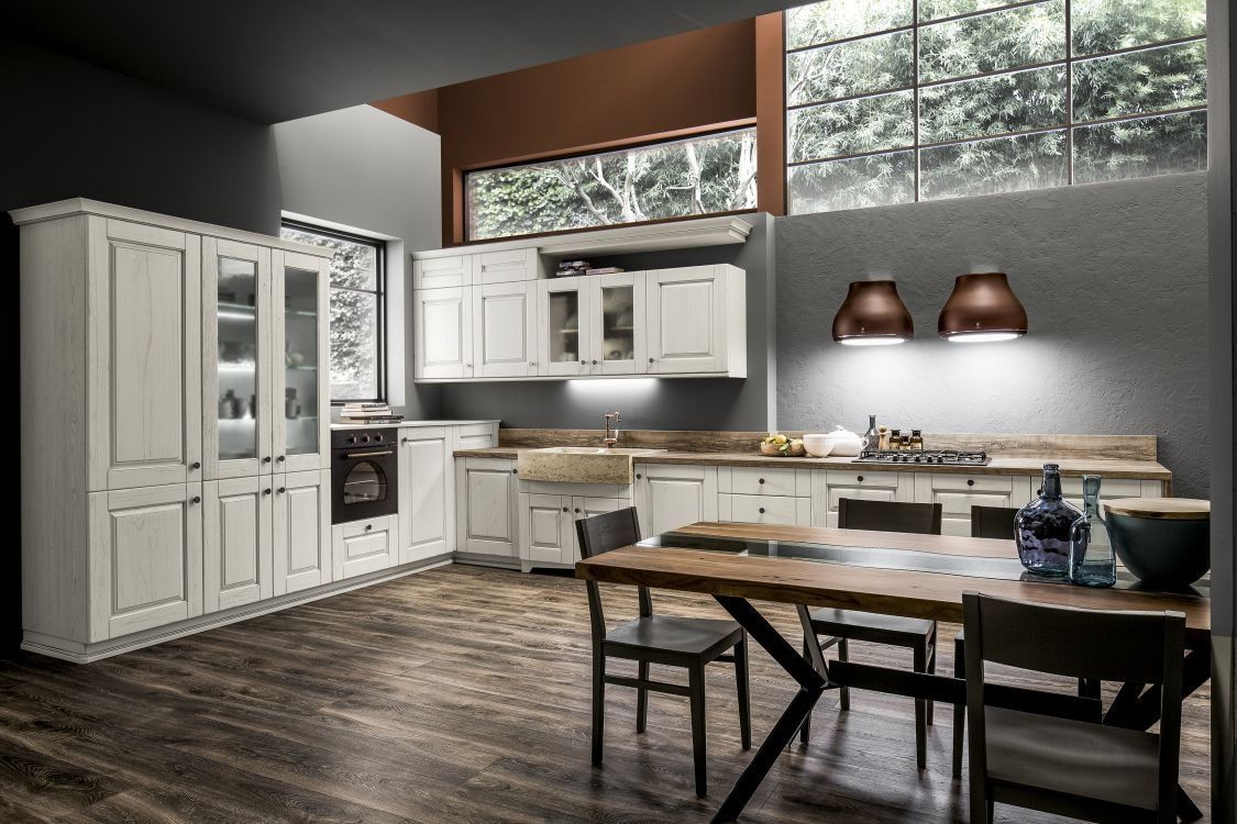 Cucina bianca con mobili in legno, ripiani e tavolo da pranzo. Stile rustico con ampie finestre e lampade a sospensione.
