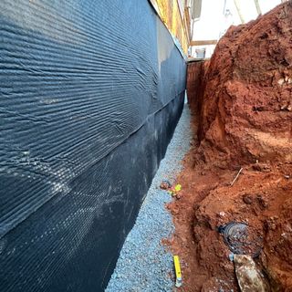 Exterior view of a foundation wall with a new waterproofing membrane installed, adjacent to an excavated area with visible drainage gravel.