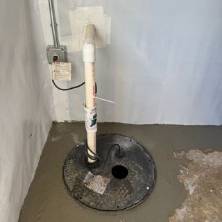 A sump pump installation in a basement, featuring a black basin with a secured lid and discharge piping against a gray wall.