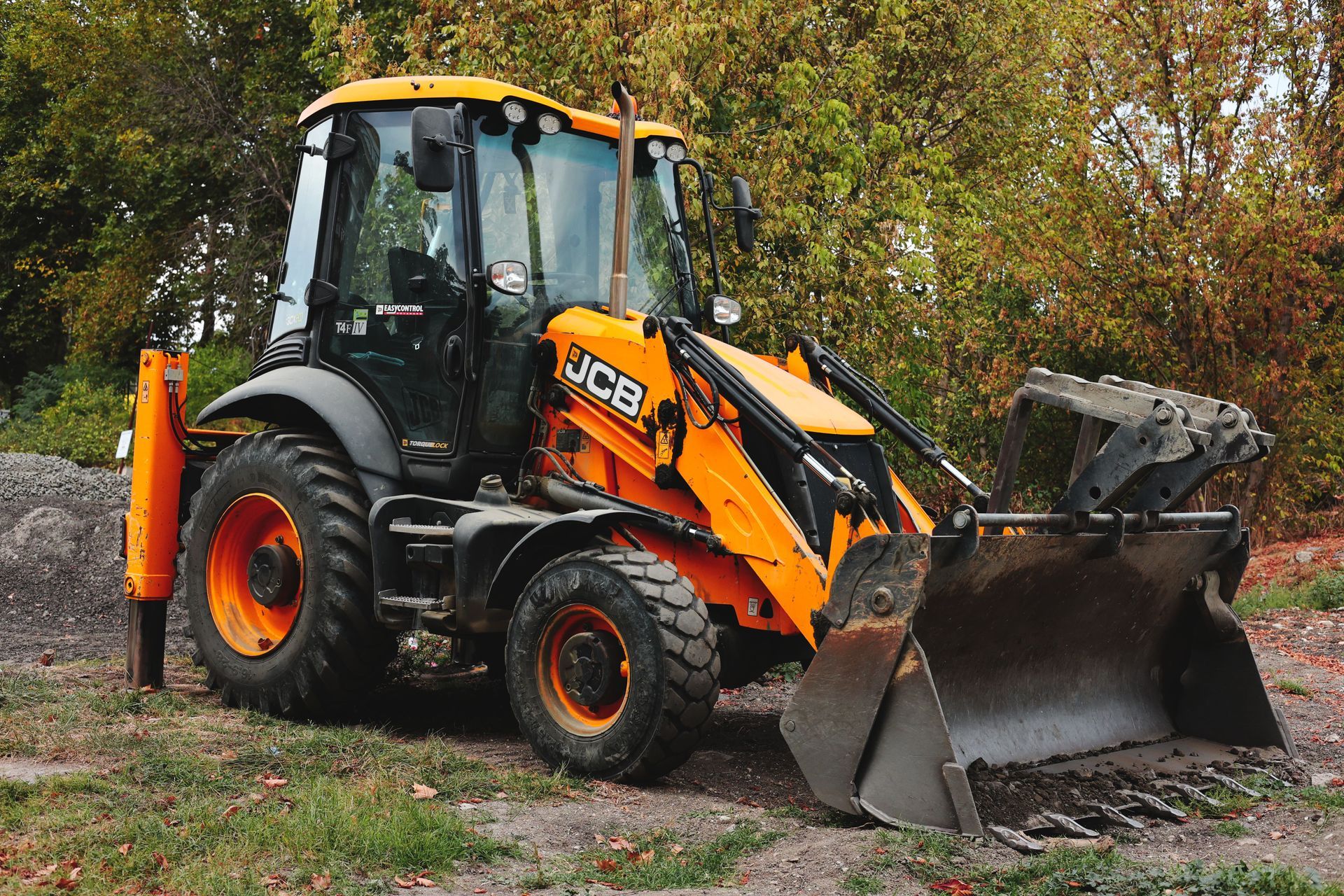 Orange JCB backhoe loader with front bucket parked on dirt in a wooded area