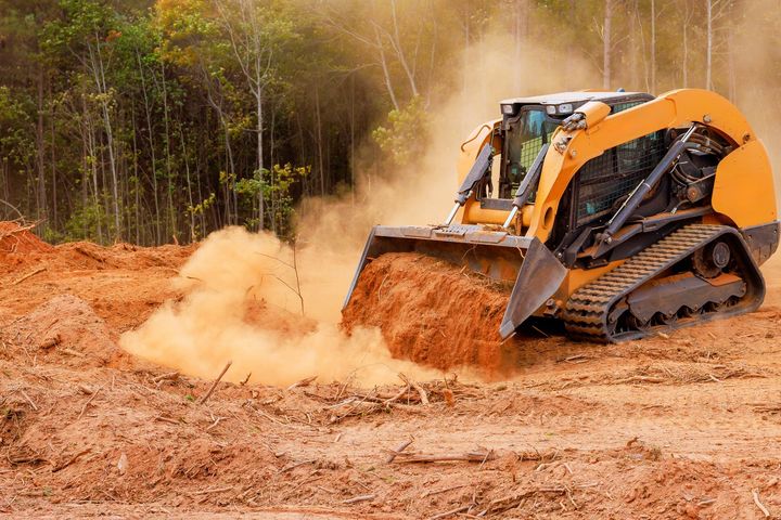 Orange skid-steer loader pushing dirt at a muddy construction site with dust cloud and trees in the background