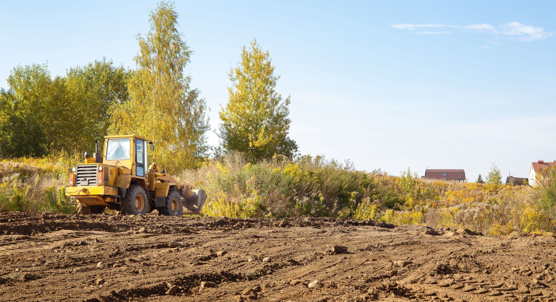 Yellow construction loader clearing a dirt field with trees and a blue sky in the background