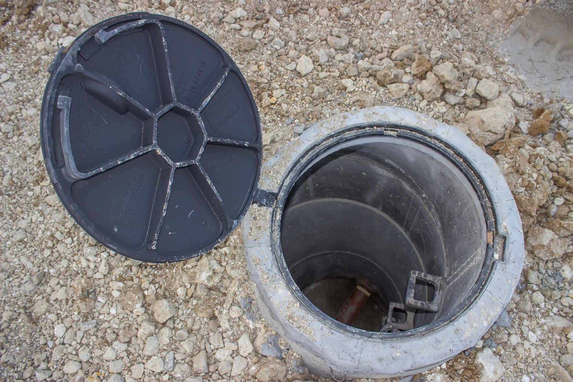 Open metal utility access hole with the black cover lid removed on gravel ground
