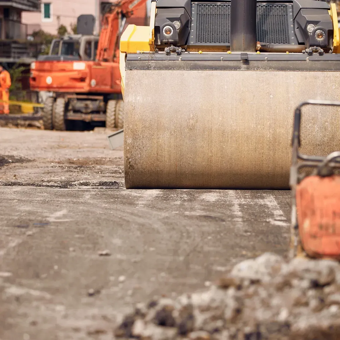 Road roller compacting dirt at a construction site, with an orange excavator in the background.
