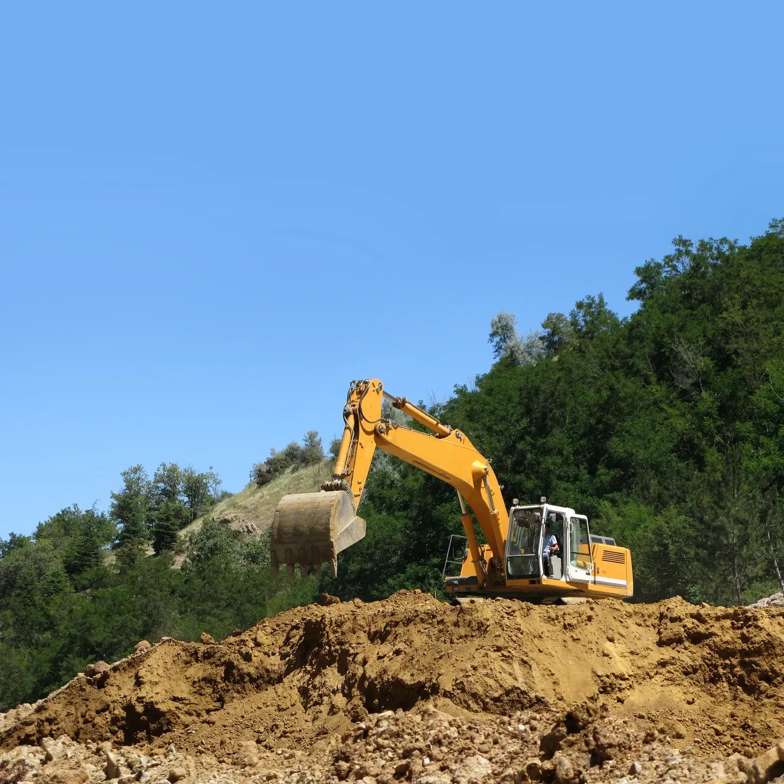 Yellow excavator working on a dirt mound beside green trees under a blue sky