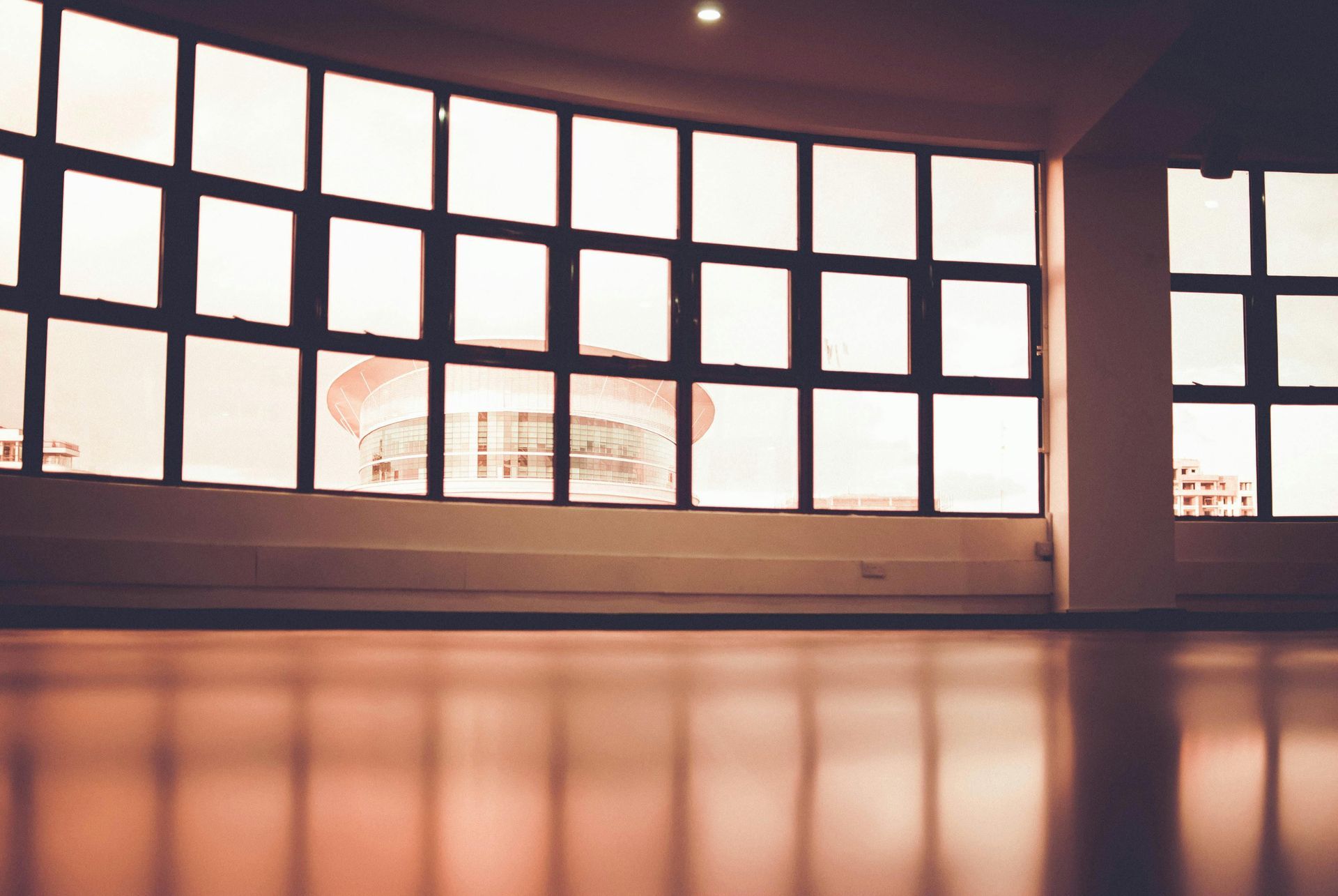 Empty room with large curved windows overlooking a distant building. Wooden floor and neutral walls.