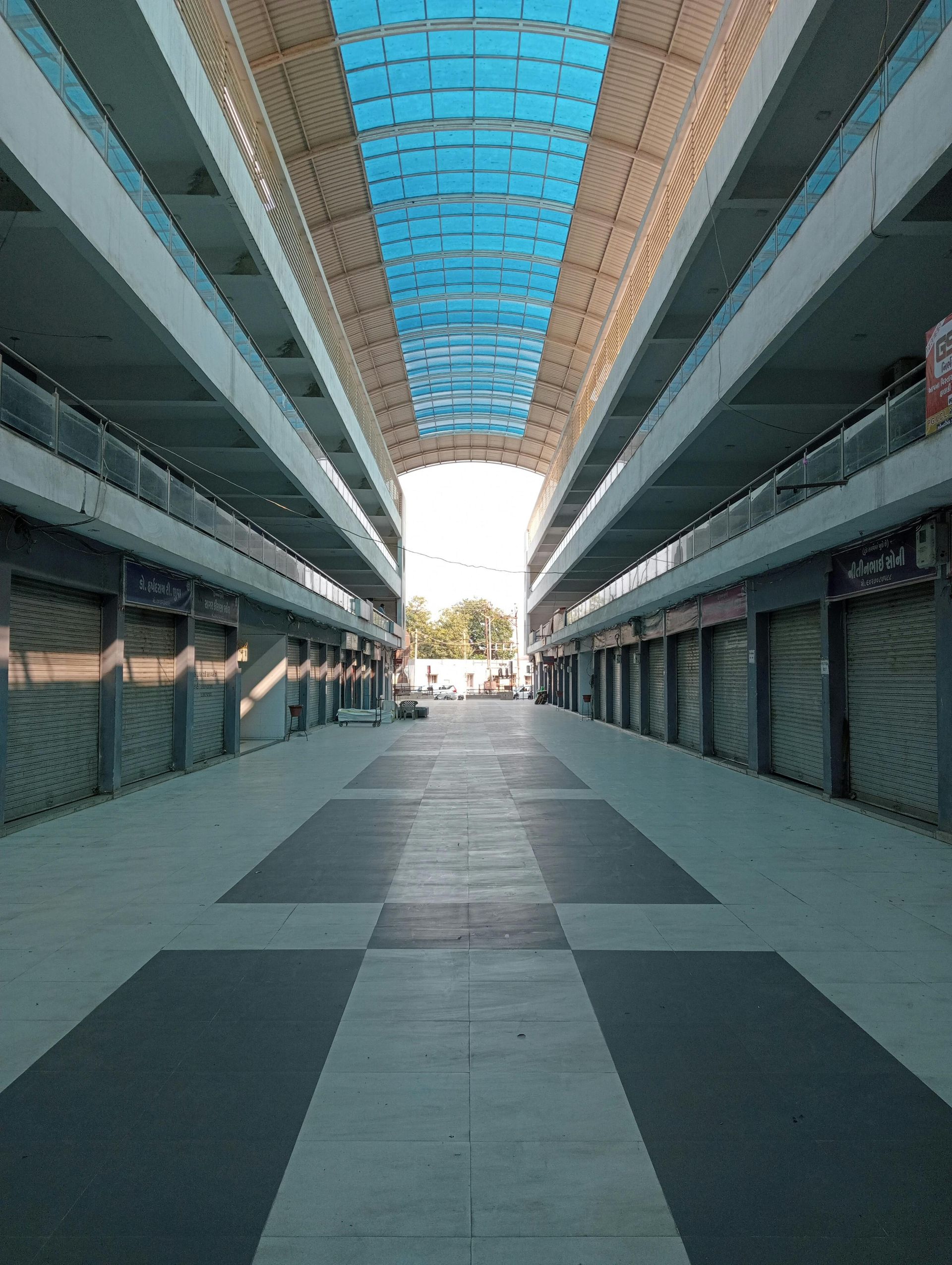 Empty shopping arcade with blue roof, rows of closed storefronts, gray and black tiled floor.