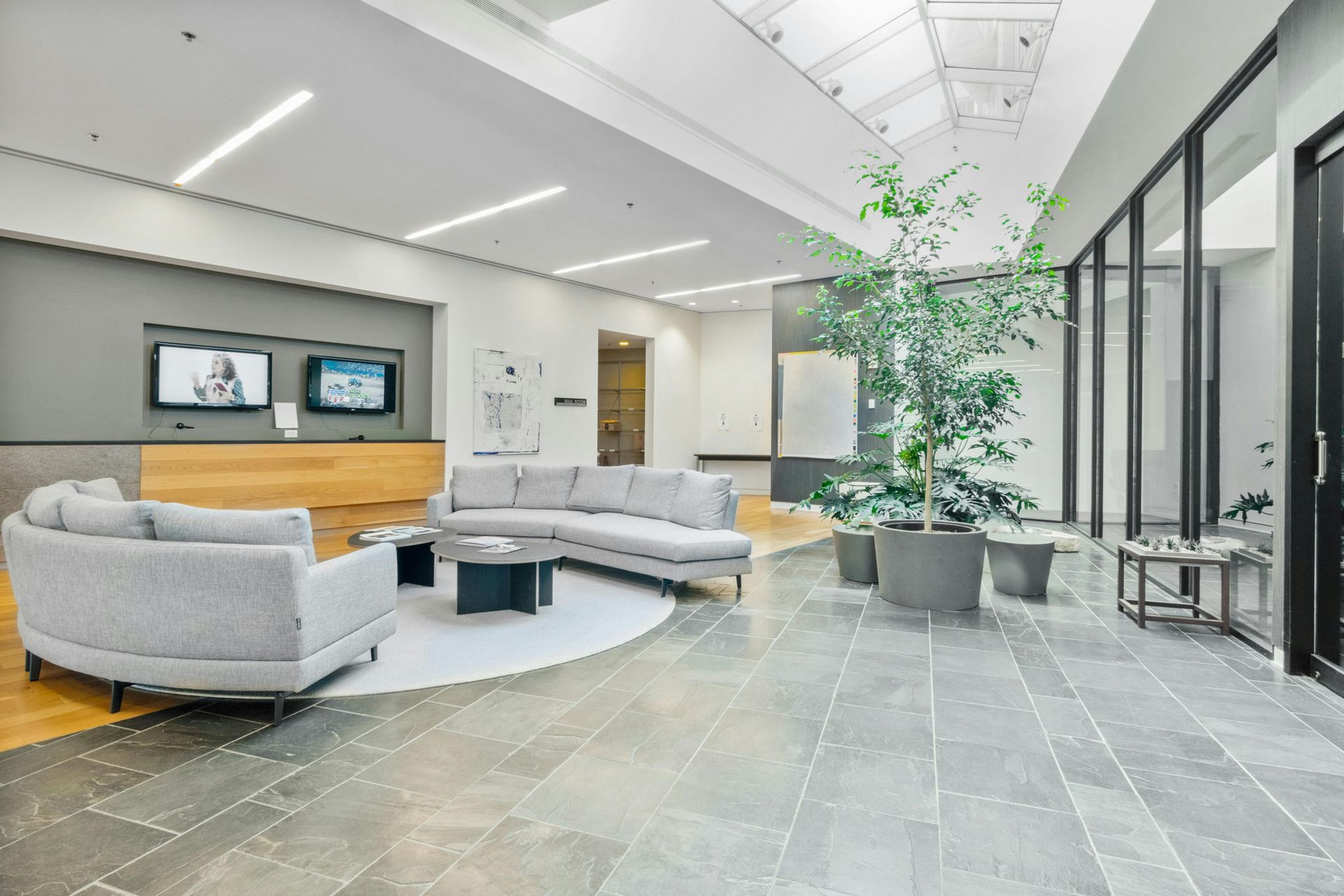 Spacious lobby with gray stone floor, curved gray sofa, large potted tree, and monitors.
