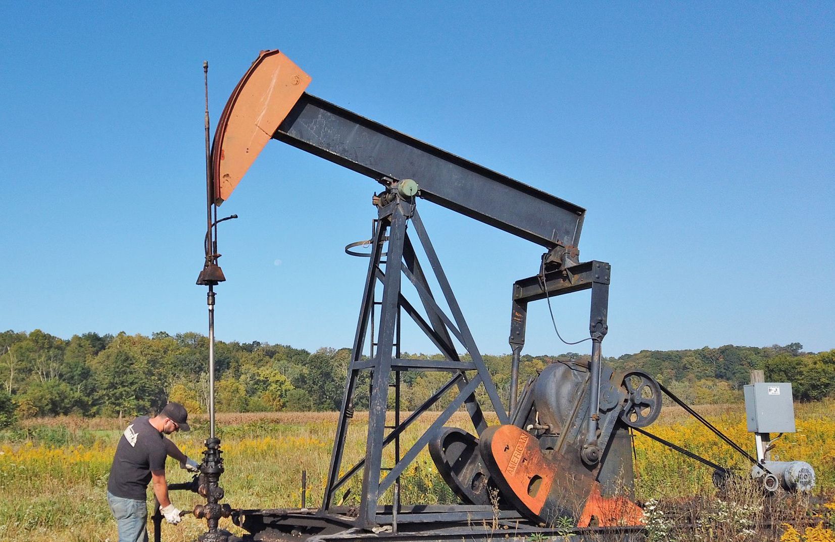 Oil pump jack in field, black and orange against blue sky, with person working on it.