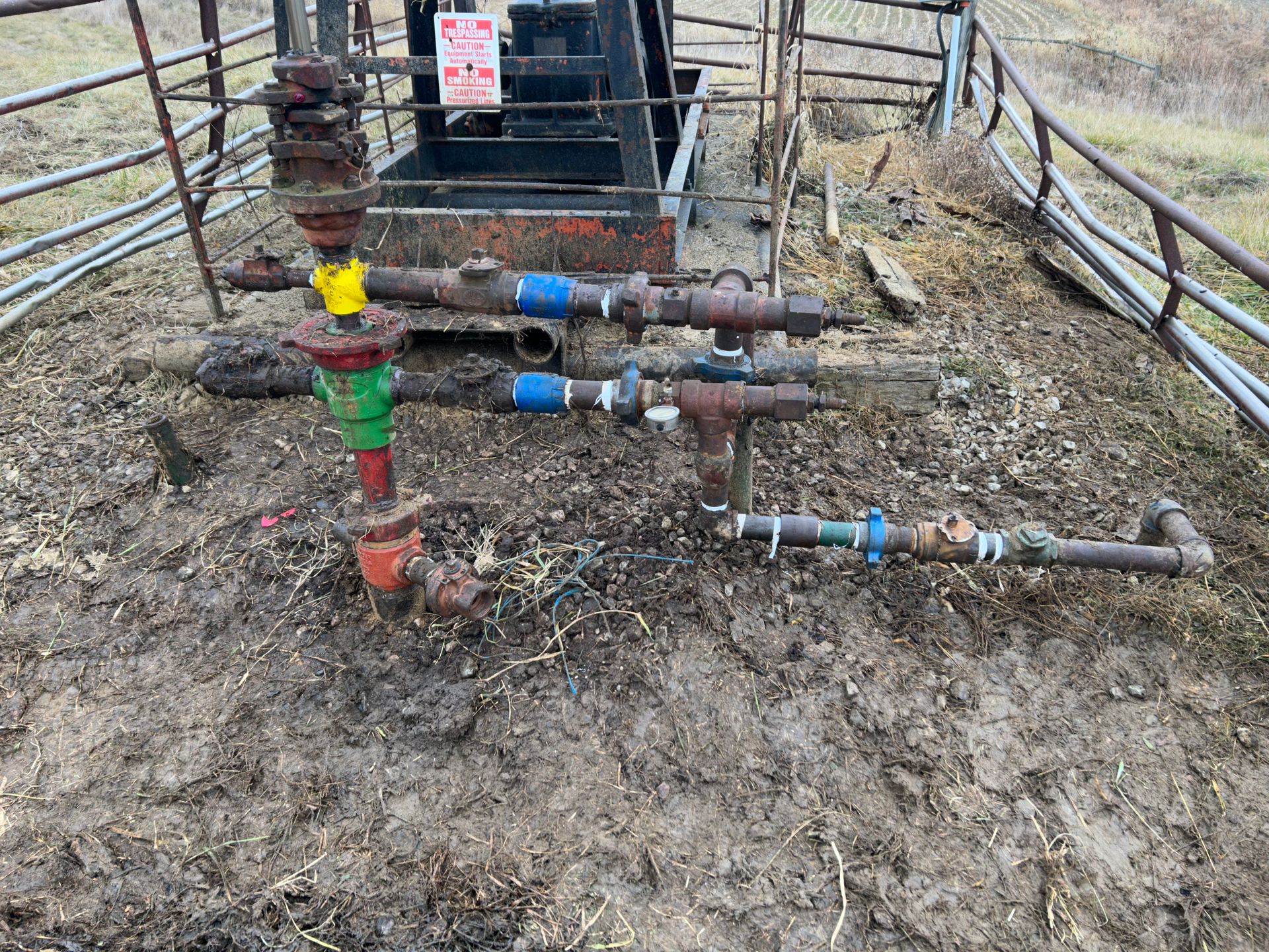 Oil well equipment with valves and pipes in a muddy outdoor setting, behind a metal fence.