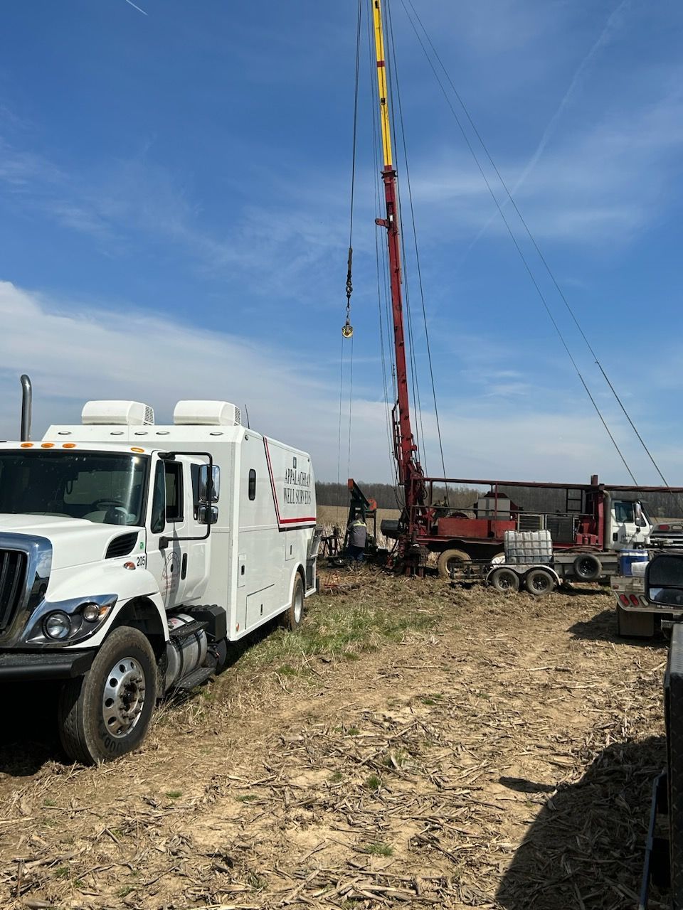 White truck and drilling rig in a field under a blue sky; worker near equipment.