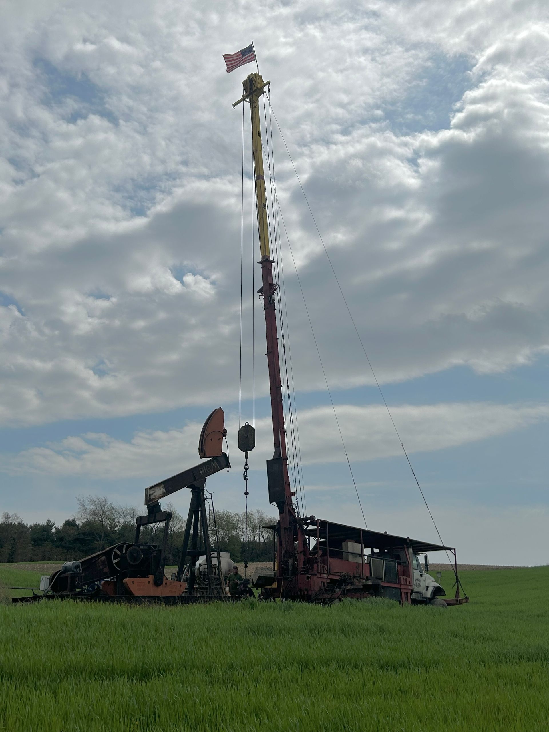 Oil rig in a green field under a cloudy sky, with a tall mast and an American flag.