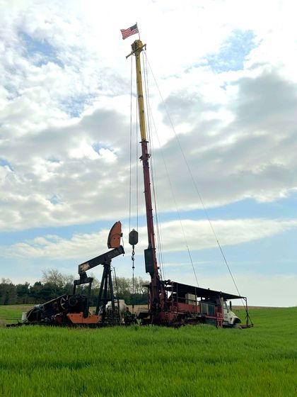 Oil derrick and pumpjack in a green field, under a cloudy sky. American flag at the top.