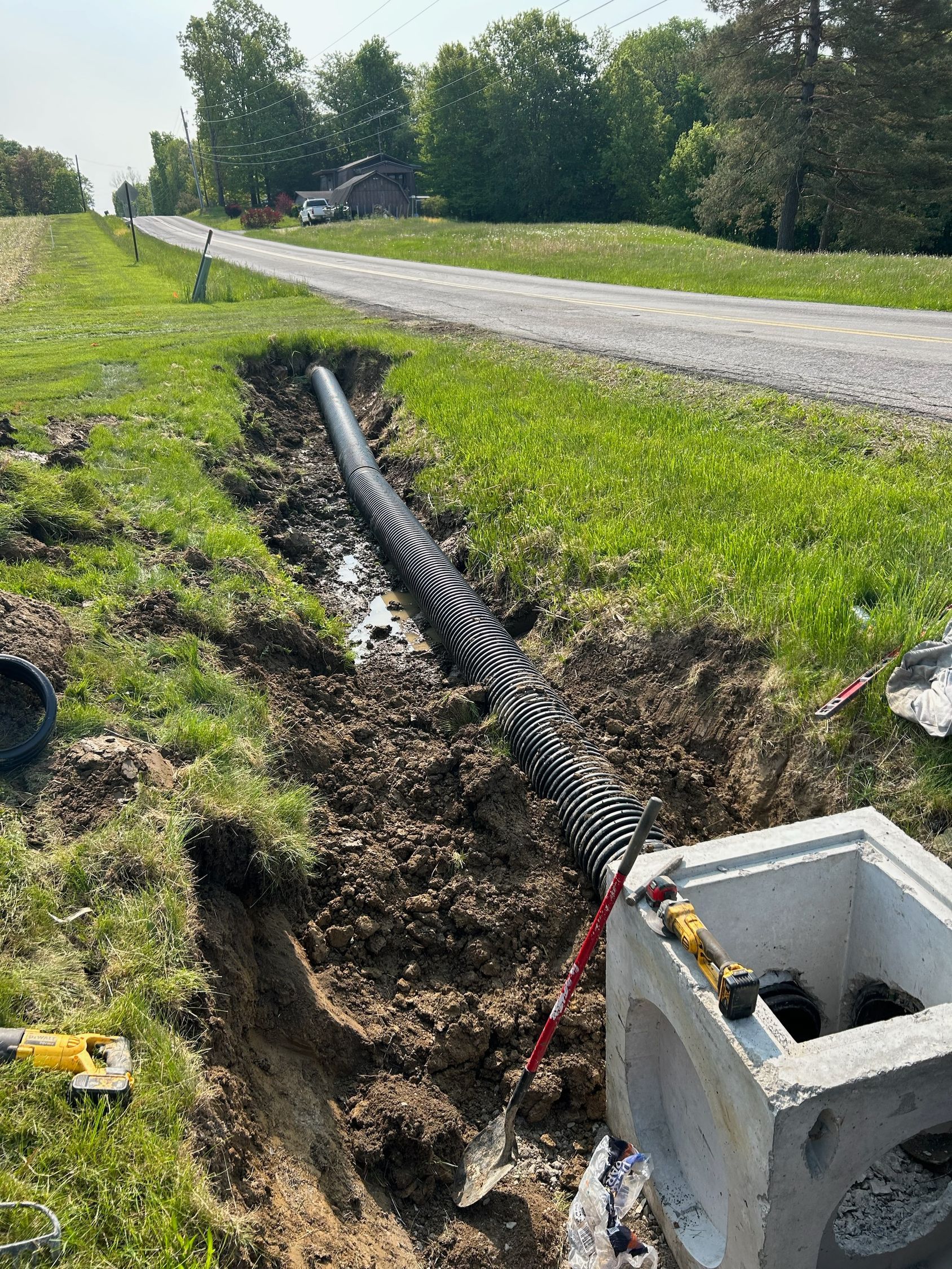 A trench with drainage pipe laid alongside a concrete structure and a gravel road in a grassy area.