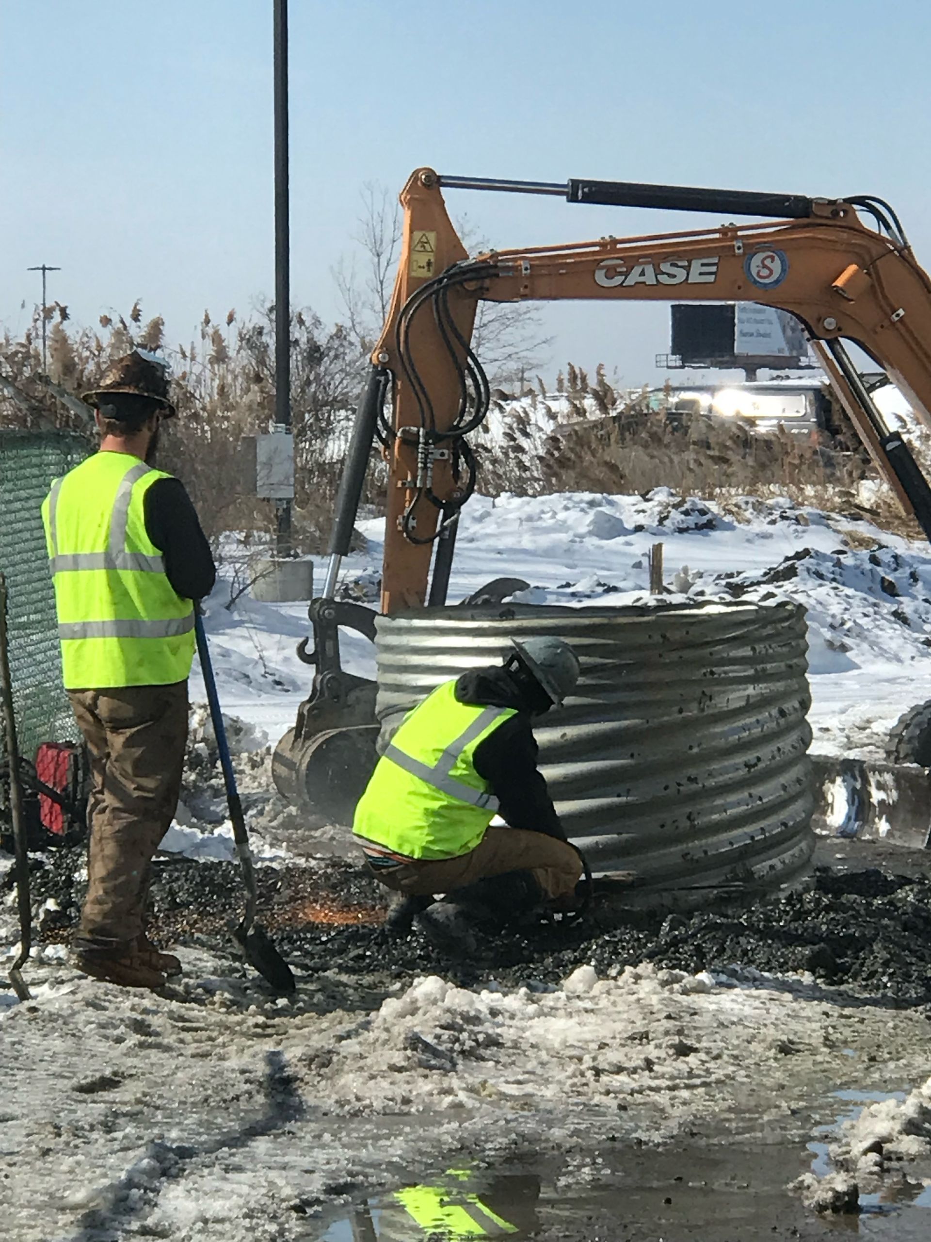 Two construction workers installing a corrugated metal pipe near an excavator; snow on the ground.