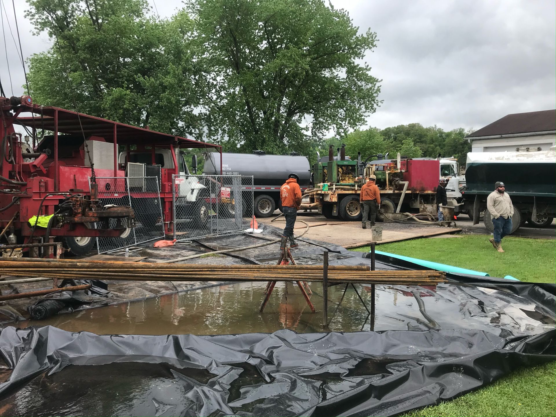 Construction site with workers and drilling equipment near a house. Puddles of water, dark tarp, and gray sky.
