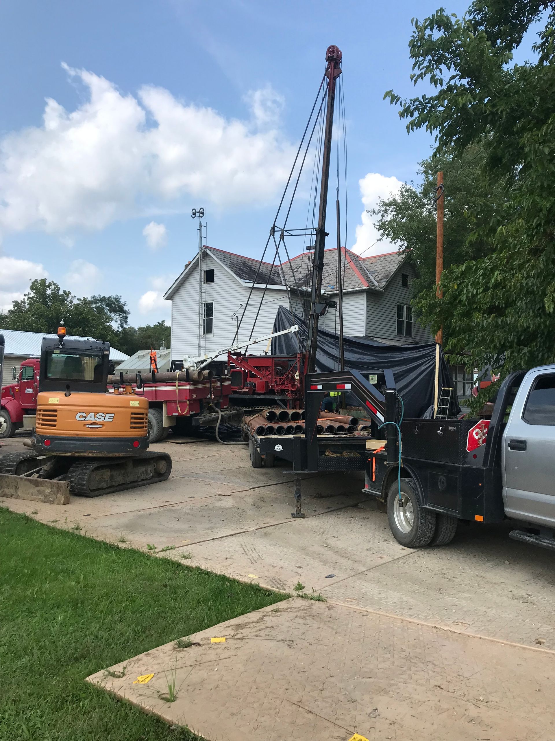 Construction site with well-drilling rig, excavator, and work trucks near a white house under a cloudy sky.