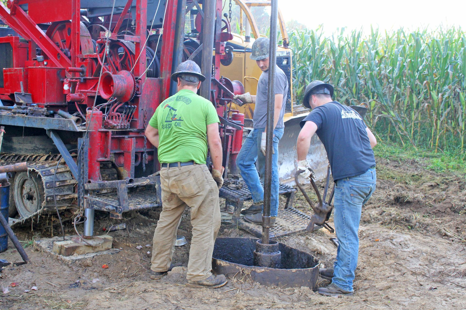 Three workers drilling a well near a cornfield. The red rig is in the background.