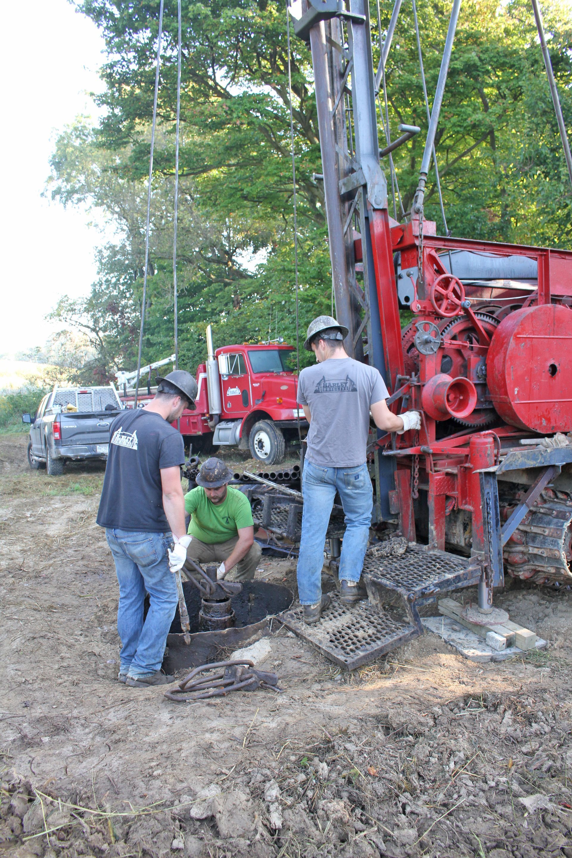Three workers operating drilling equipment on muddy ground near a red truck.