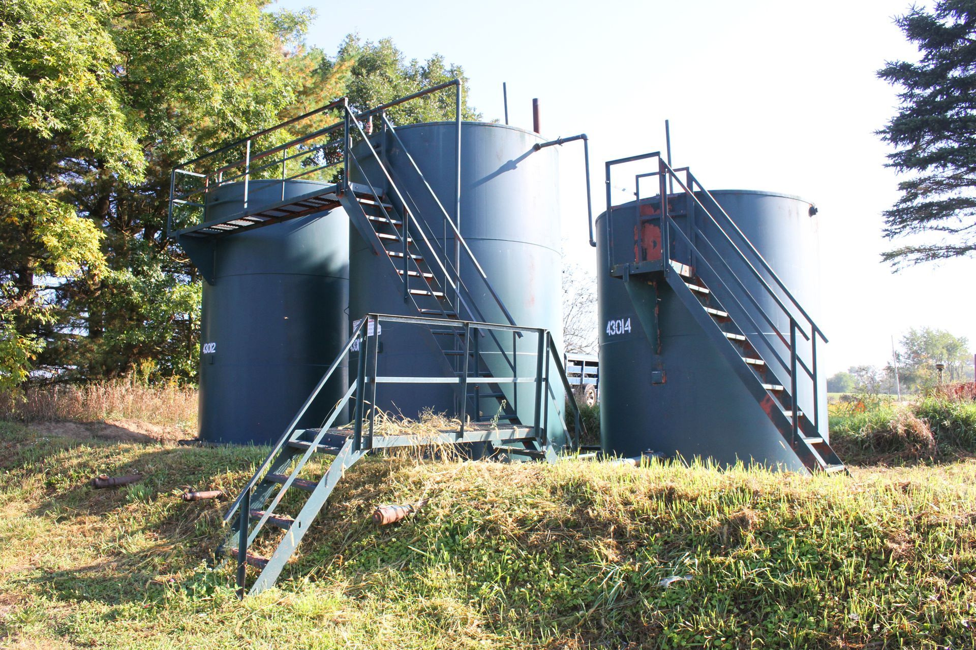 Three dark green industrial tanks with metal staircases, set outdoors with grass and trees.