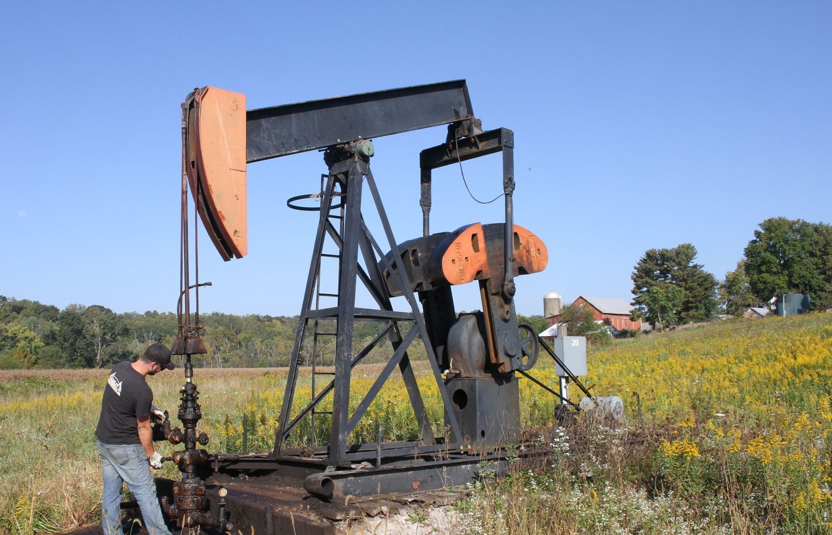Man working on a black and orange oil pump in a field of wildflowers under a blue sky.
